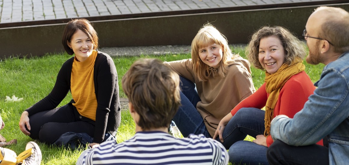 Viisi ihmistä istuu nurmikolla auringonpaisteessa keskustelemassa