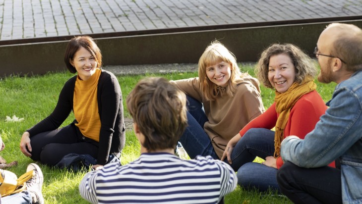 Viisi ihmistä istuu nurmikolla auringonpaisteessa keskustelemassa