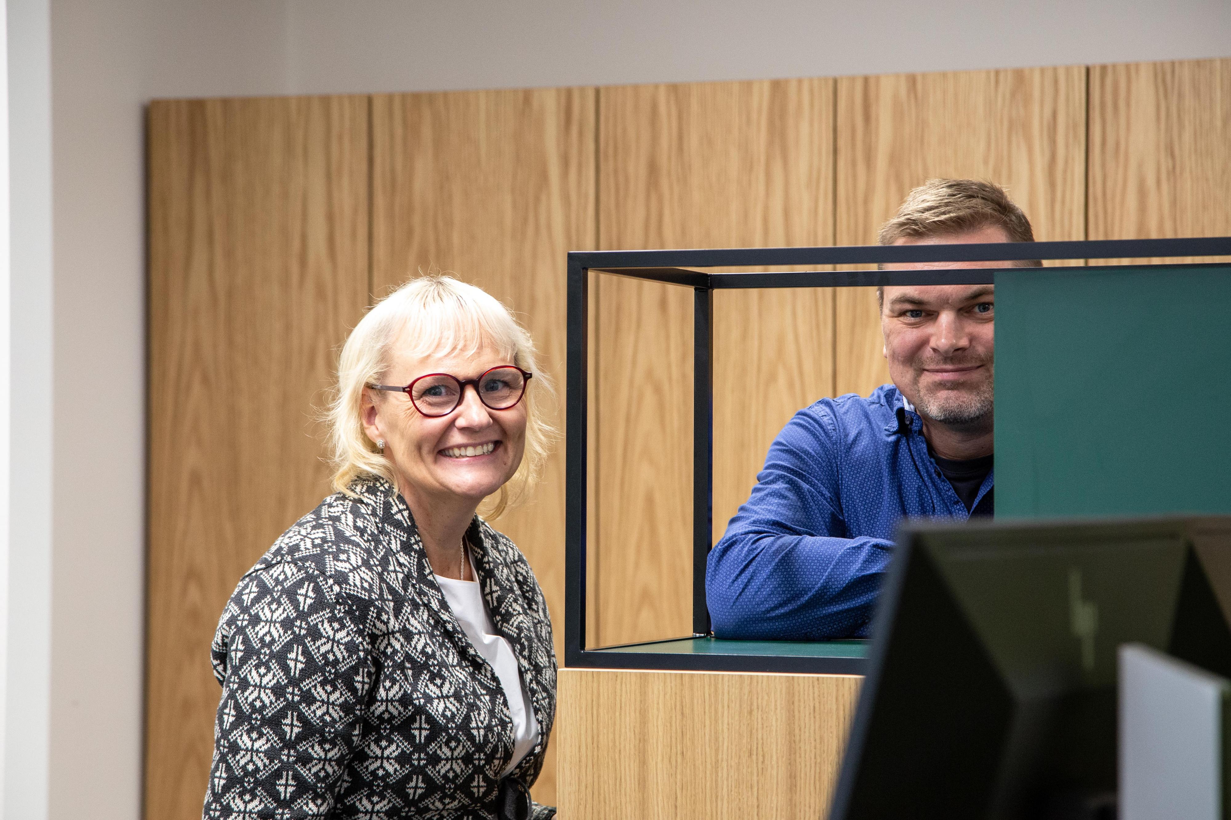 Nainen ja mies seisvat toimistohyllyjen vieressä ja katsovat kameraan iloisesti nauraen