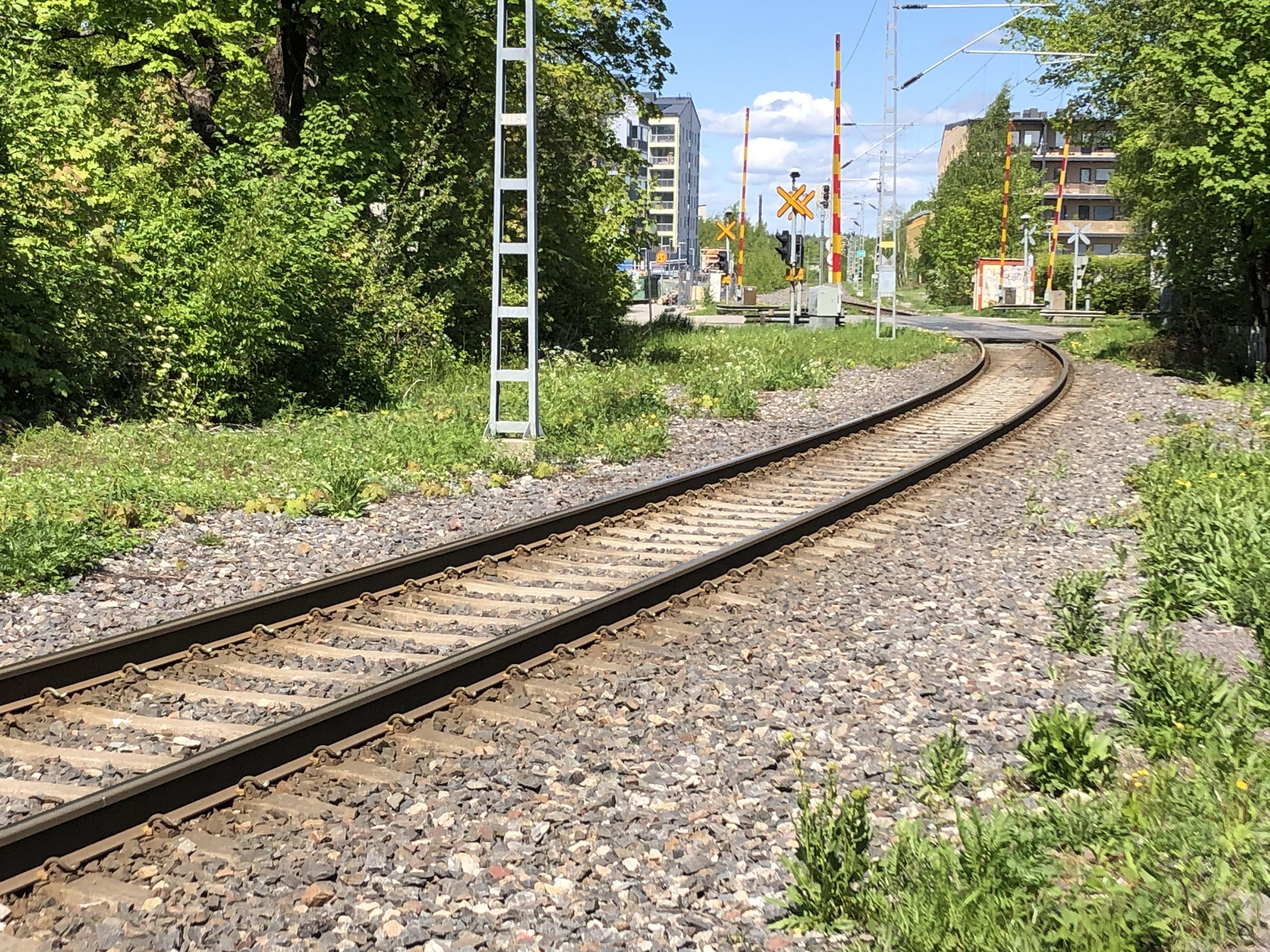 Puiden ja pensaiden keskellä kulkevat junaraiteet, taustalla kerrostaloja ja tasoristeys