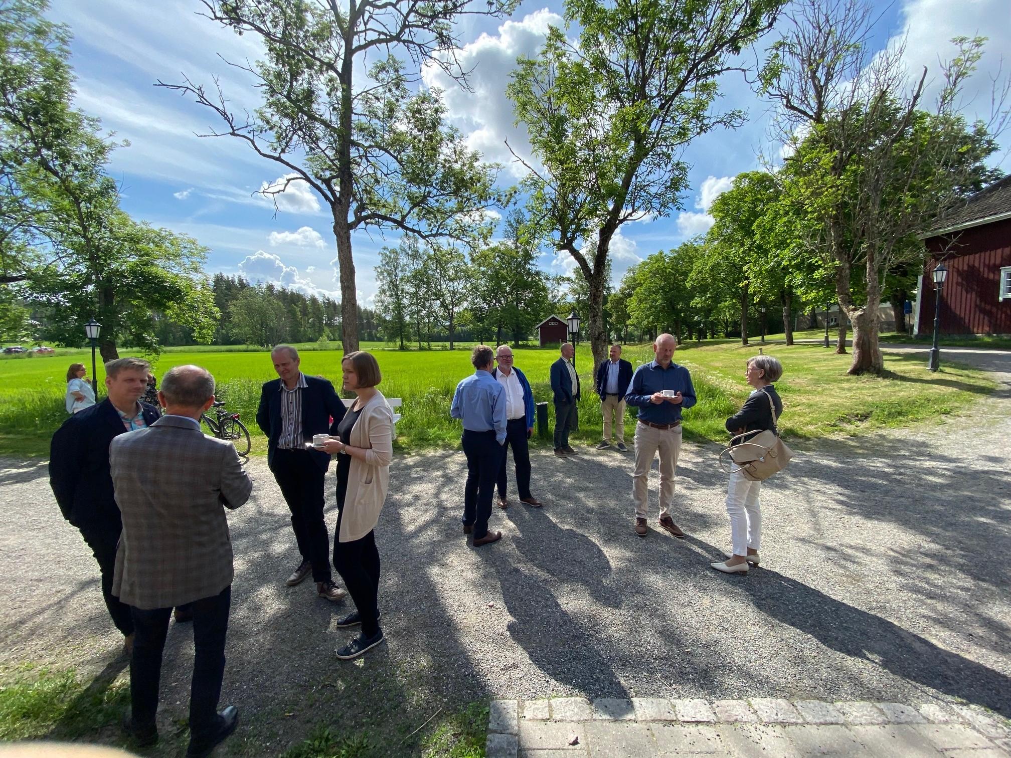 Joukko naisia ja miehiä seisoo viheriöivällä pihalla keskustelemassa vilkkaasti