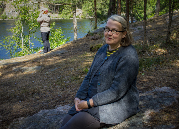 Metsäisessä mäessä hymyilevä Jaana Itälä-Laine istumassa maassa, taustalla vettä ja henkilö puhumassa puhelimessa. 