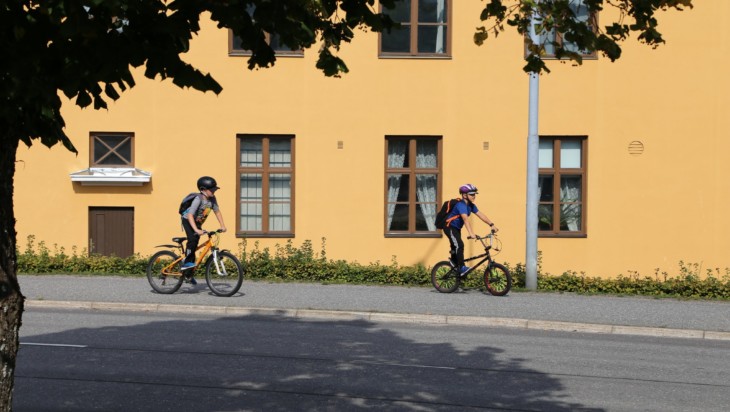 Keltaisen kivitalon edustalla kaksi koululaista pyöräilemässä t-paitasillaan kypärät päässään.