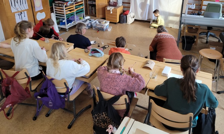 Neljä oppilasta istuu pulpeteissaan piirtämässä. Oppilaita ja opettajia istuu myös lattialla taiteilemassa. Luokan edessä istuu mallina yksi oppilas.