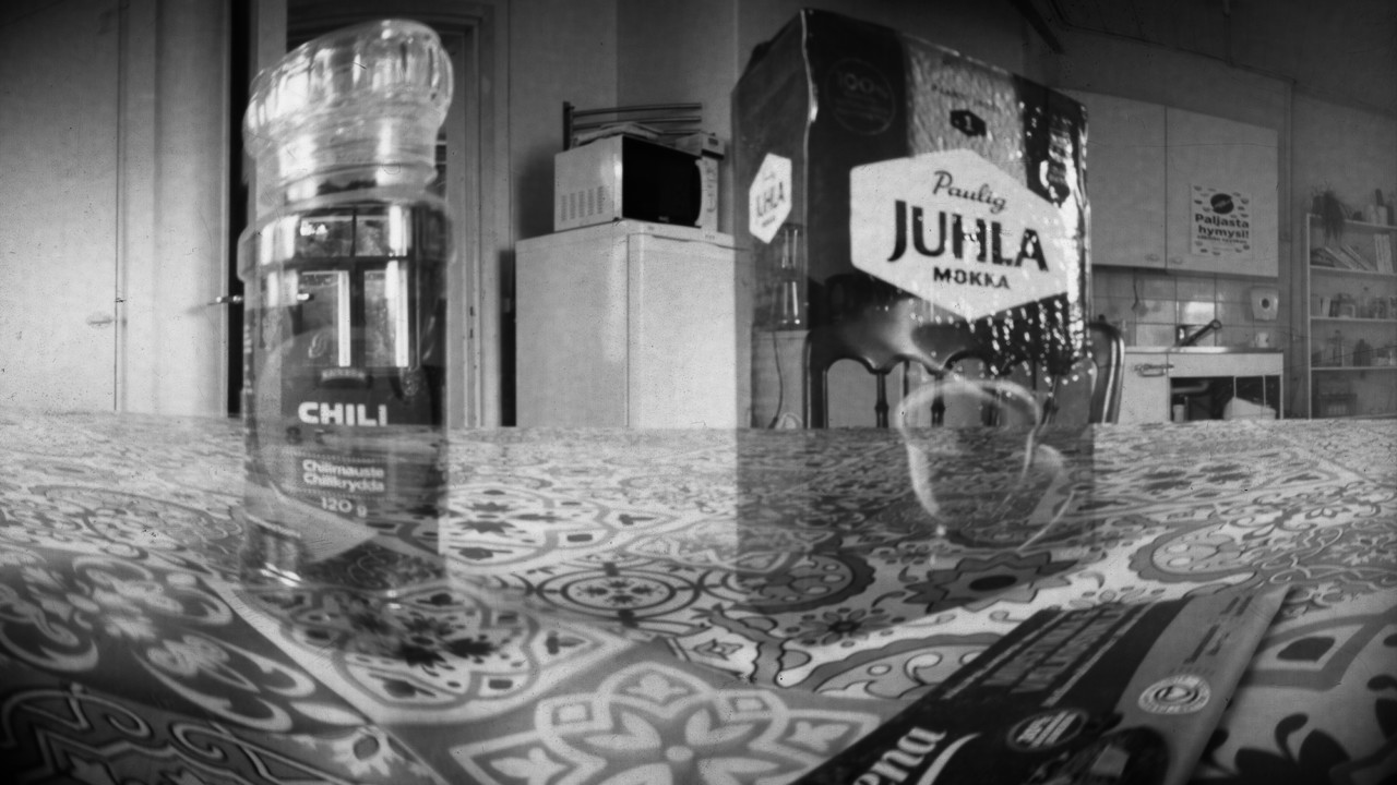 Camera obscura -tekniikalla otettu mustavalkoinen kuva. Kuvassa näkyy taustalla keittiö, etualalla liinalla peitetyllä pöydällä mm. Juhalmokka-paketti..