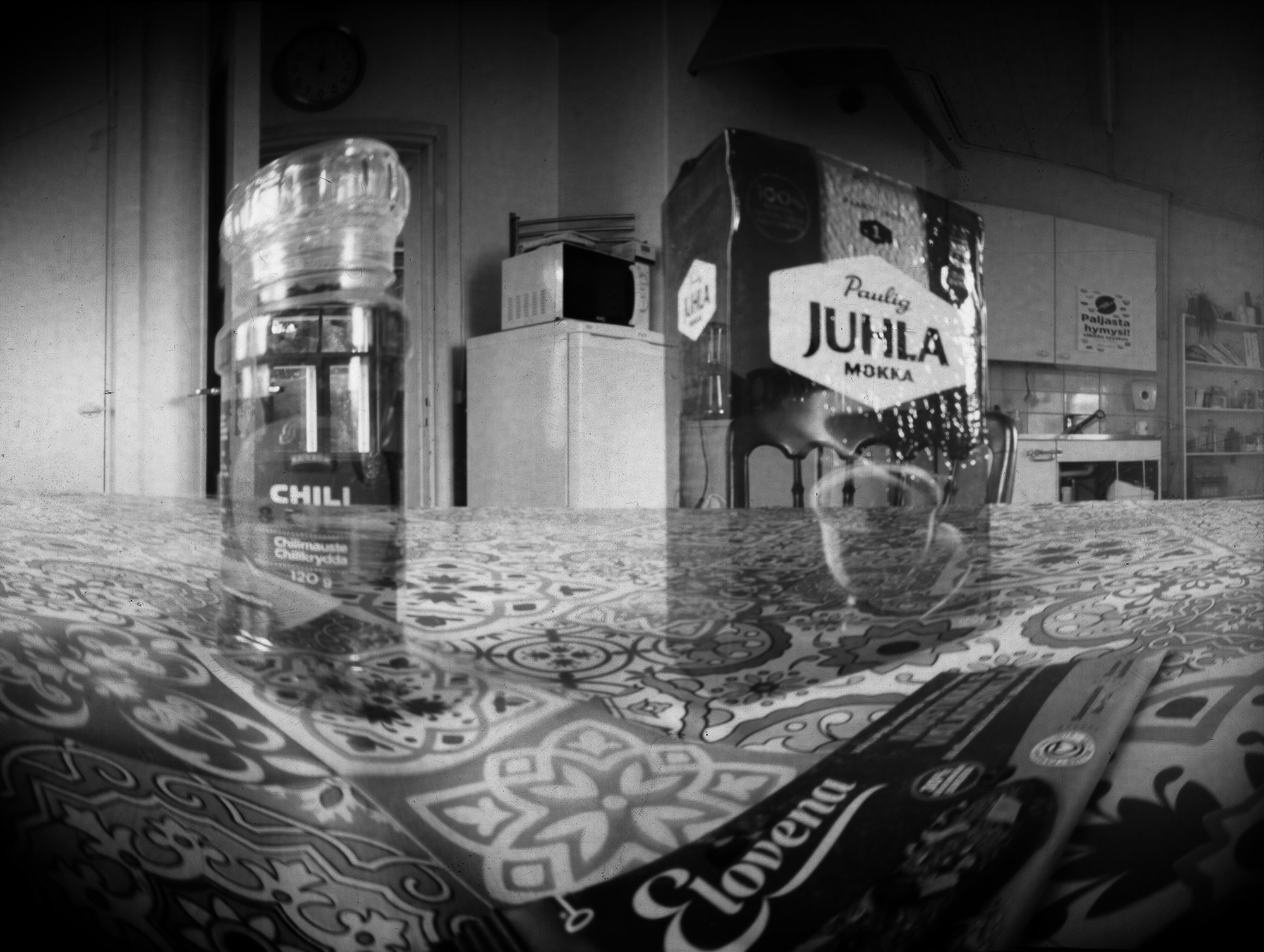 Camera obscura -tekniikalla otettu mustavalkoinen kuva. Kuvassa näkyy taustalla keittiö, etualalla liinalla peitetyllä pöydällä mm. Juhalmokka-paketti..