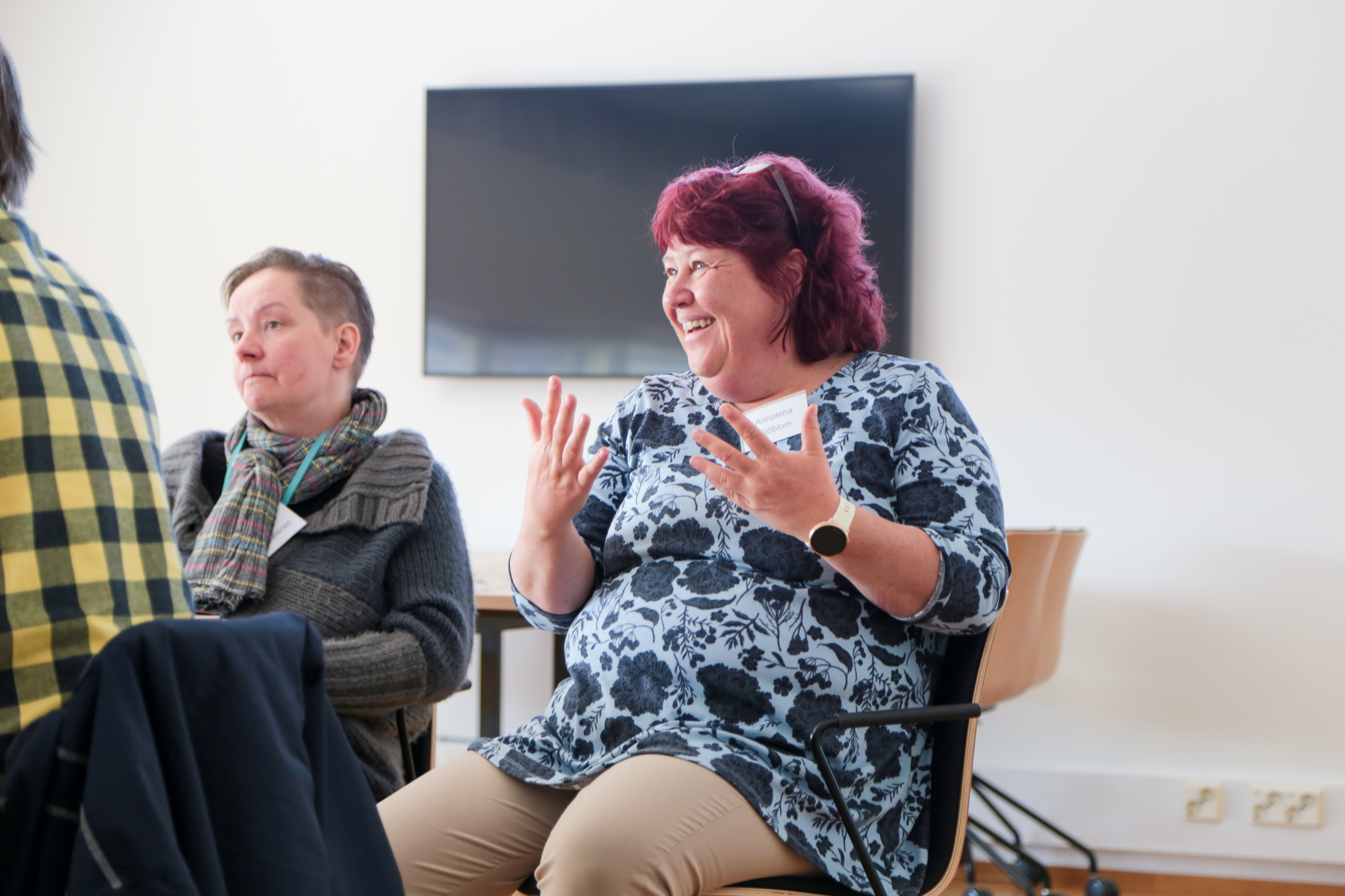 Naisoletettu hymyilee ja liikuttaa käsiään puheenvuoron tehostamiseksi. Toinen henkilö kuuntelee vieressä.