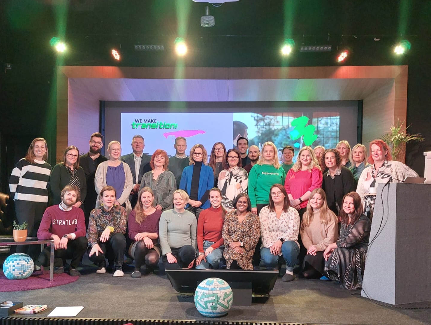 Kuva menetelmäharjoituksen osallistujista, noin 35 henkilöstä lavalla ison hankelogon edessä.