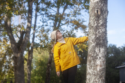 Amanda Vuori pukeutuneena keltaiseen takkiin ja beigeen hattuun. Hän pitää vasemmalla kädellään kiinni puun rungosta ja katsoo ylös kohti puun latvaa.