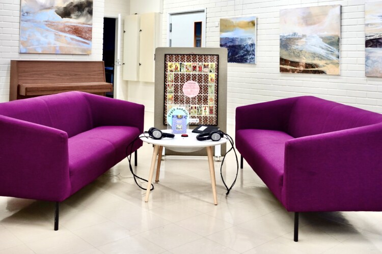 Kaksi toisiaan vastapäätä olevaa violettia sohvaa vaaleassa huoneessa. Sohvien välissä on pyöreä pöytä, jolla kahdet kuulokkeet ääni-installaation kuuntelua varten.