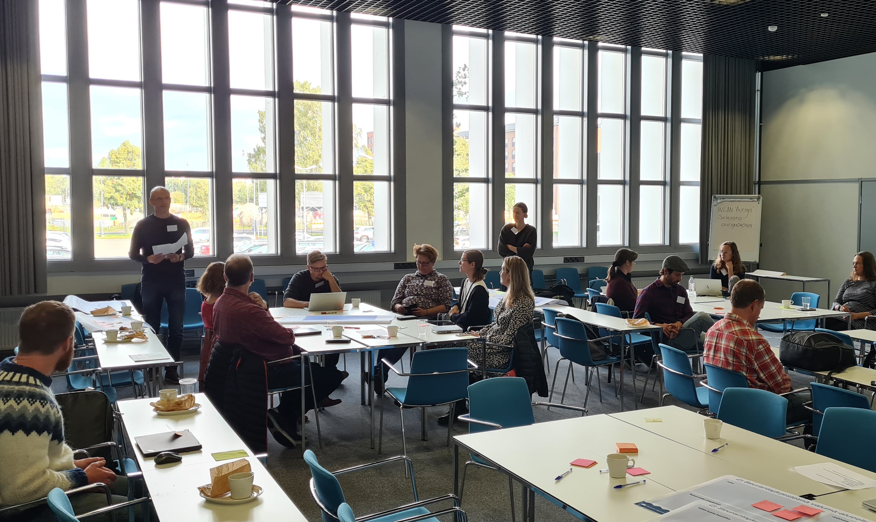 Ihmisiä istuu työpajassa isossa salissa pöytien ääressä keskustelemassa