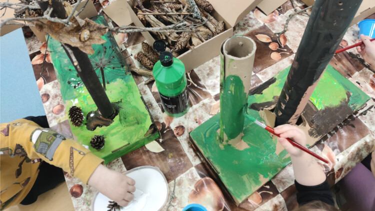Pöydällä on mm. pahvirullia, käpyjä ja oksia sekä liimaa ja maaleja. Kuvassa näkyy kahden lapsen kädet. He liimaavat materiaaleja yhteen ja maalaavat pahvirullia vihreiksi.