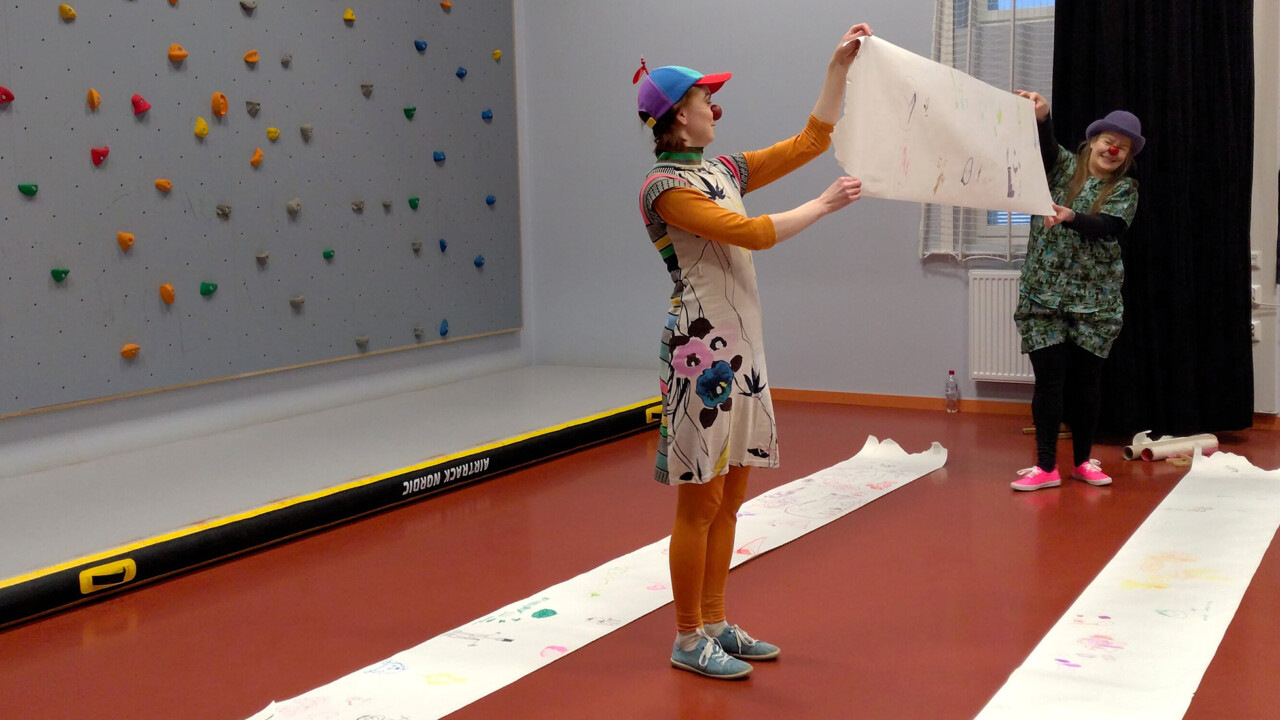 Kaksi värikkäästi pukeutunutta klovnia pitää välissään paperia, johon on piirretty jotakin. Taustalla koulun jumppasalin kiipeilyseinä.