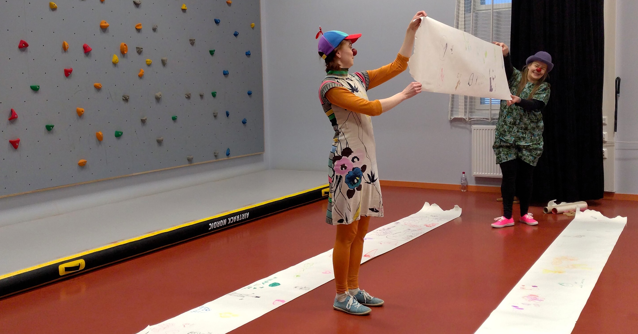 Kaksi värikkäästi pukeutunutta klovnia pitää välissään paperia, johon on piirretty jotakin. Taustalla koulun jumppasalin kiipeilyseinä.