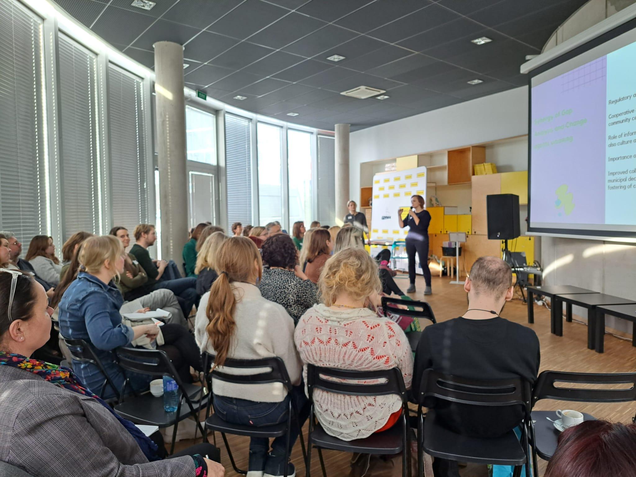 Esitystilanne, jossa yksi henkilö puhuu noin 50 hengen yleisölle diaesityksen kanssa.