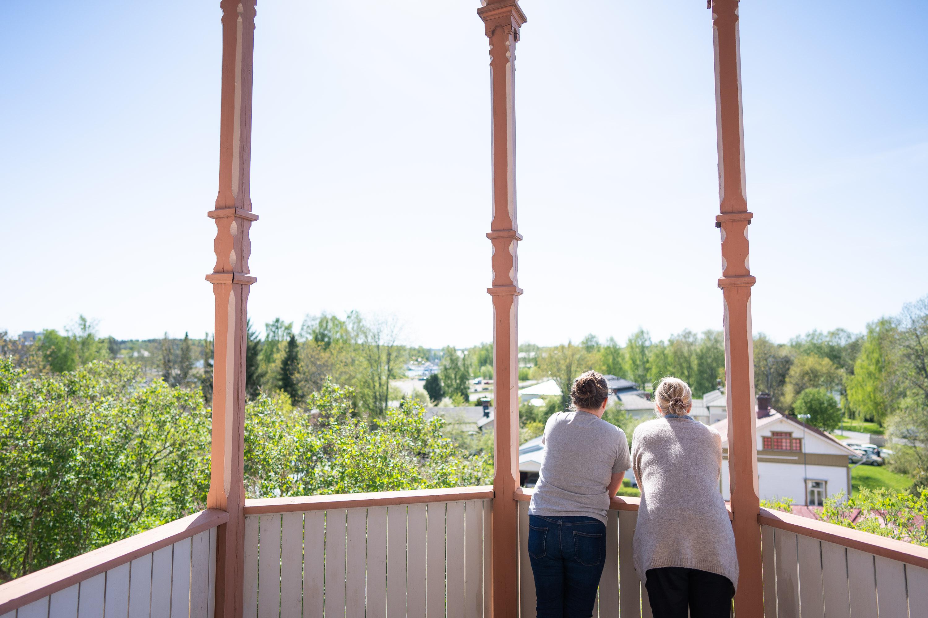 Kaksi ihmistä selkä päin kameraan katsovat rakennuksen terassilta alas vihreään maisemaan.