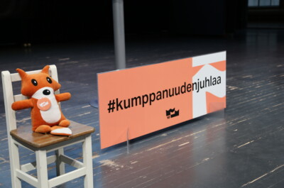 Kettupehmo istuu puisella tuolilla. Vieressä on oranssi kyltti jossa lukee Kumppanuuden juhlaa.