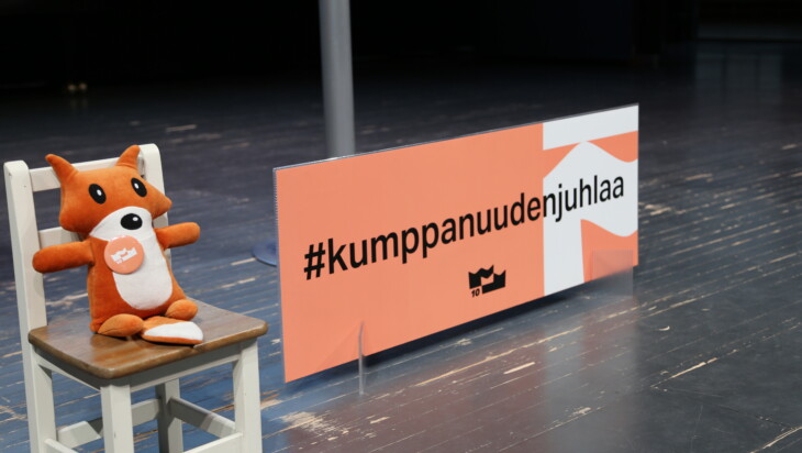 Kettupehmo istuu puisella tuolilla. Vieressä on oranssi kyltti jossa lukee Kumppanuuden juhlaa.