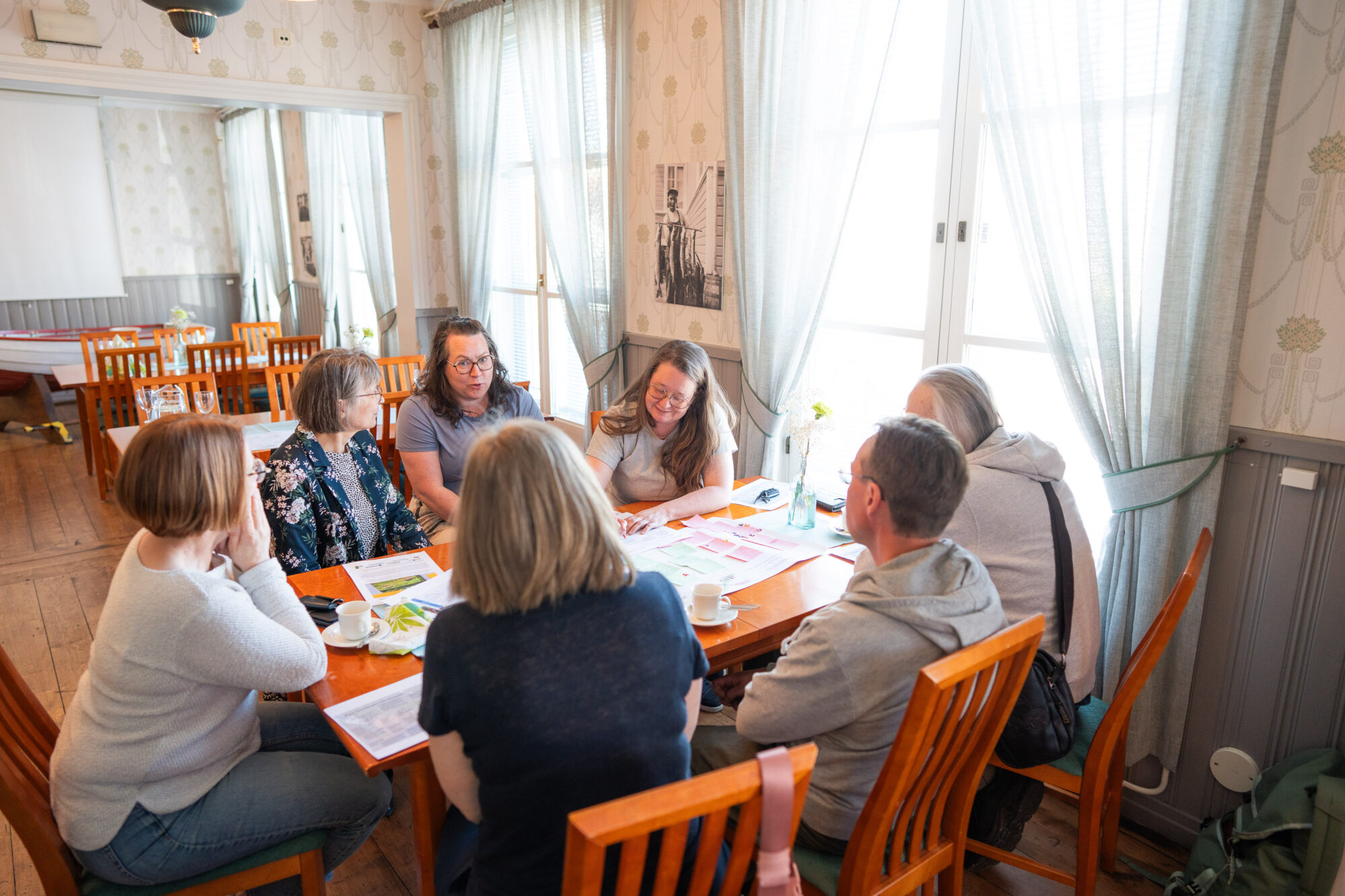 Ihmisiä pöydän ääressä. Pöydällä on papereita, joissa on post-it-lappuja. Valoisa tunnelma.