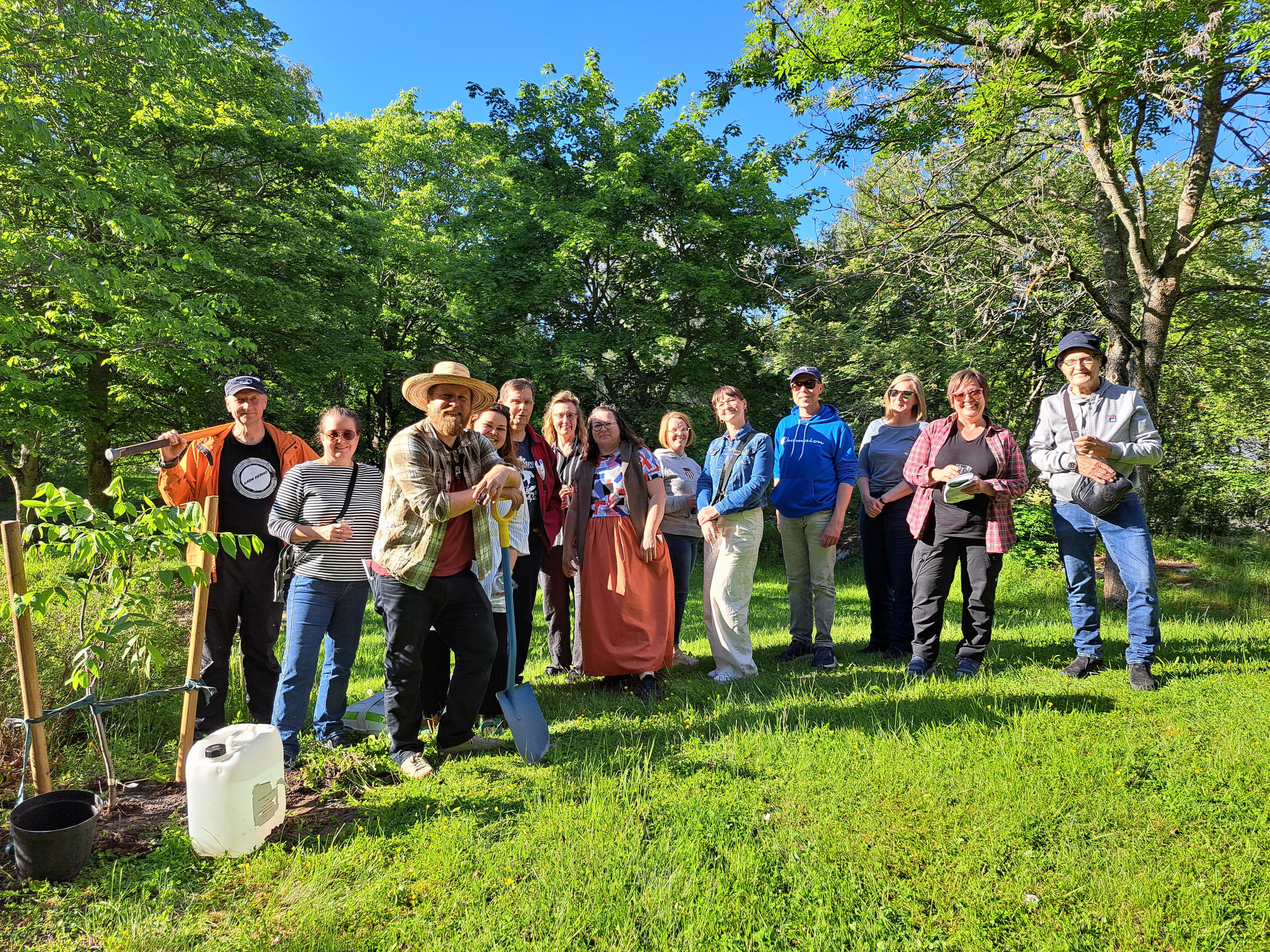 Noin 15 hymyilevää ihmistä vehreässä ympäristössä aurinkoisella säällä. Yhdellä on rautalapio.