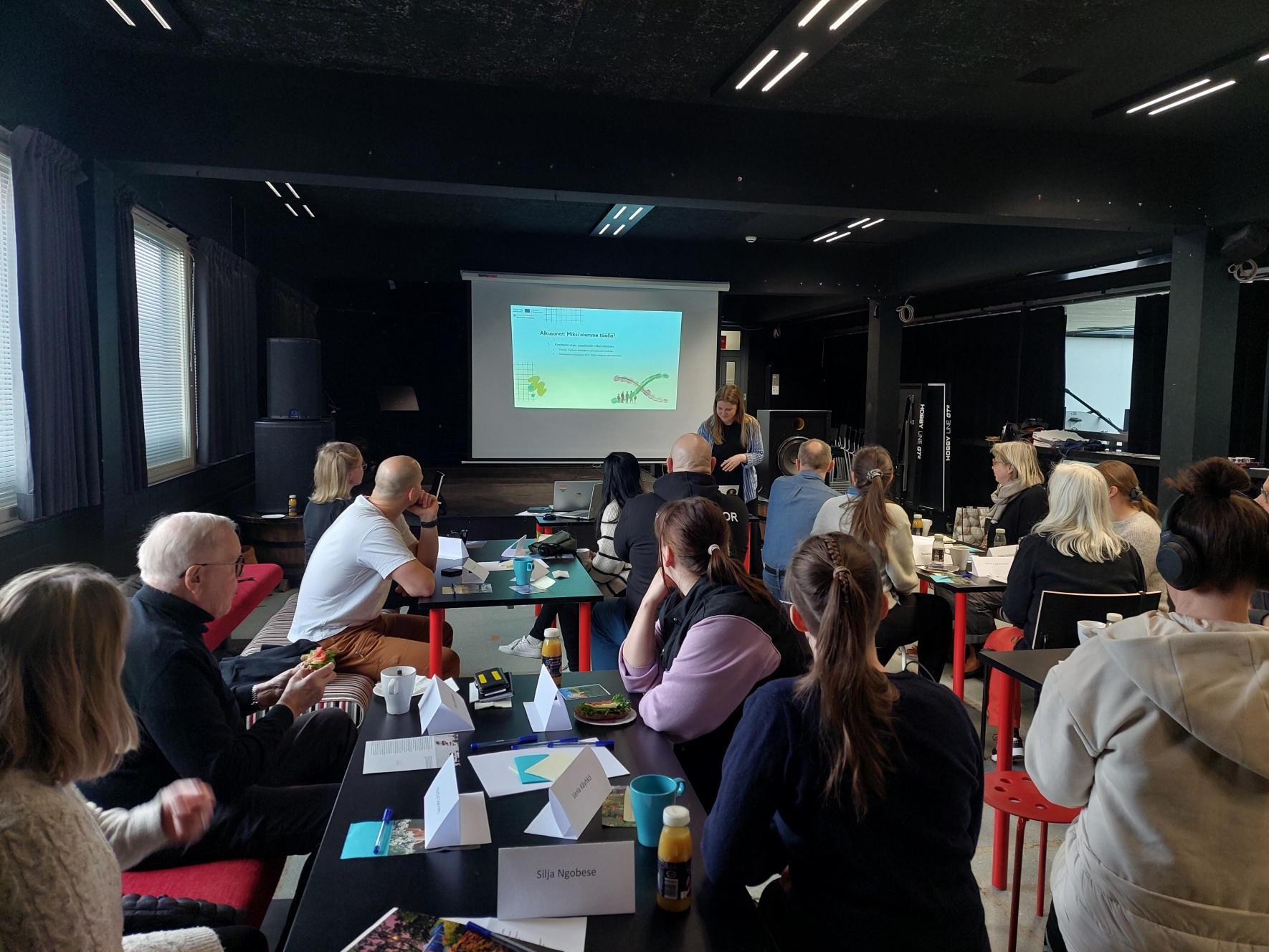 Kuva työpajatilanteesta. Edessä diaesitys ja esittäjä, yleisö istuu pöytäryhmittäin.