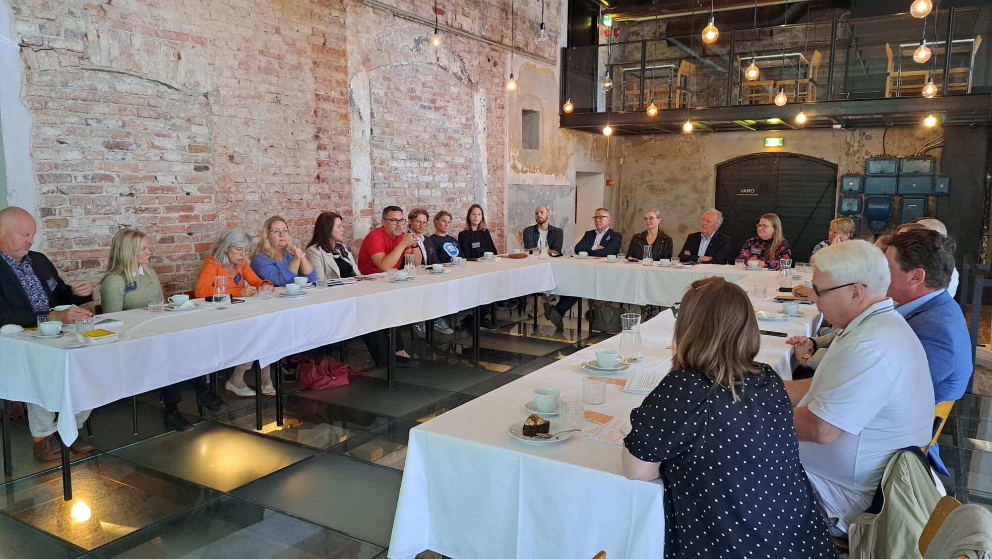 Noin 20 ihmistä U:n muotoisen pöydän ääressä keskustelemassa.