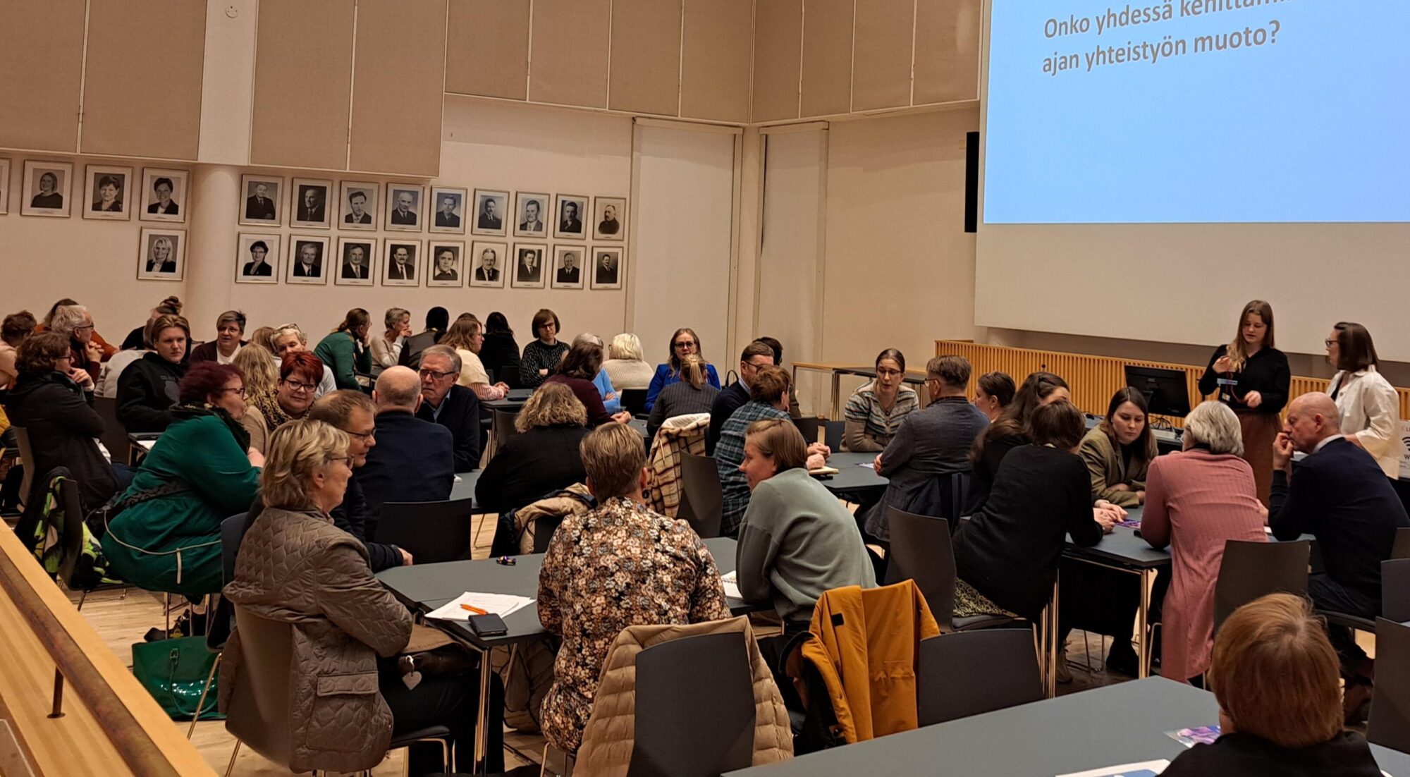 Ihmisiä istumassa auditorion etuosassa samalla tasolla olevissa 4-5 hengen pöydissä. Diaesitys taustalla.