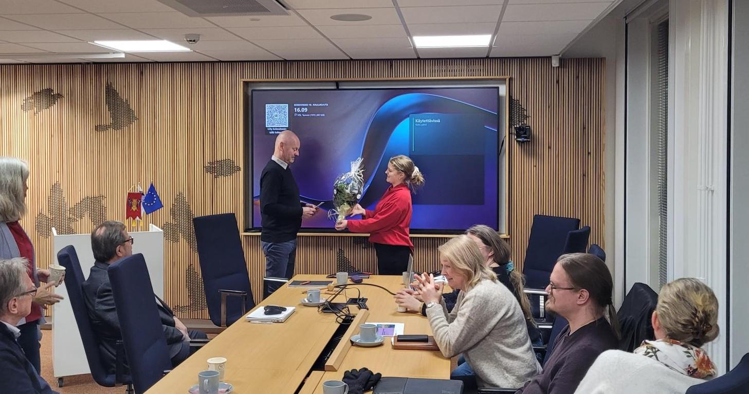 Yksi henkilö ojentaa toiselle kukkakimpun. Pöydän ääressä istuvat ihmiset katsovat.