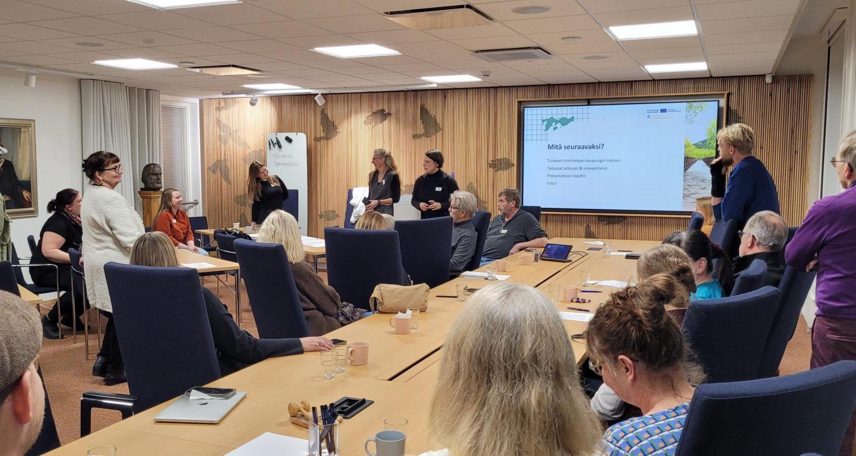 Kaksi ihmistä esiintyy ja muut katsovat. Huone, jossa on iso pöytä, valkokangas ja paljon puupintoja. Noin 20 ihmistä.