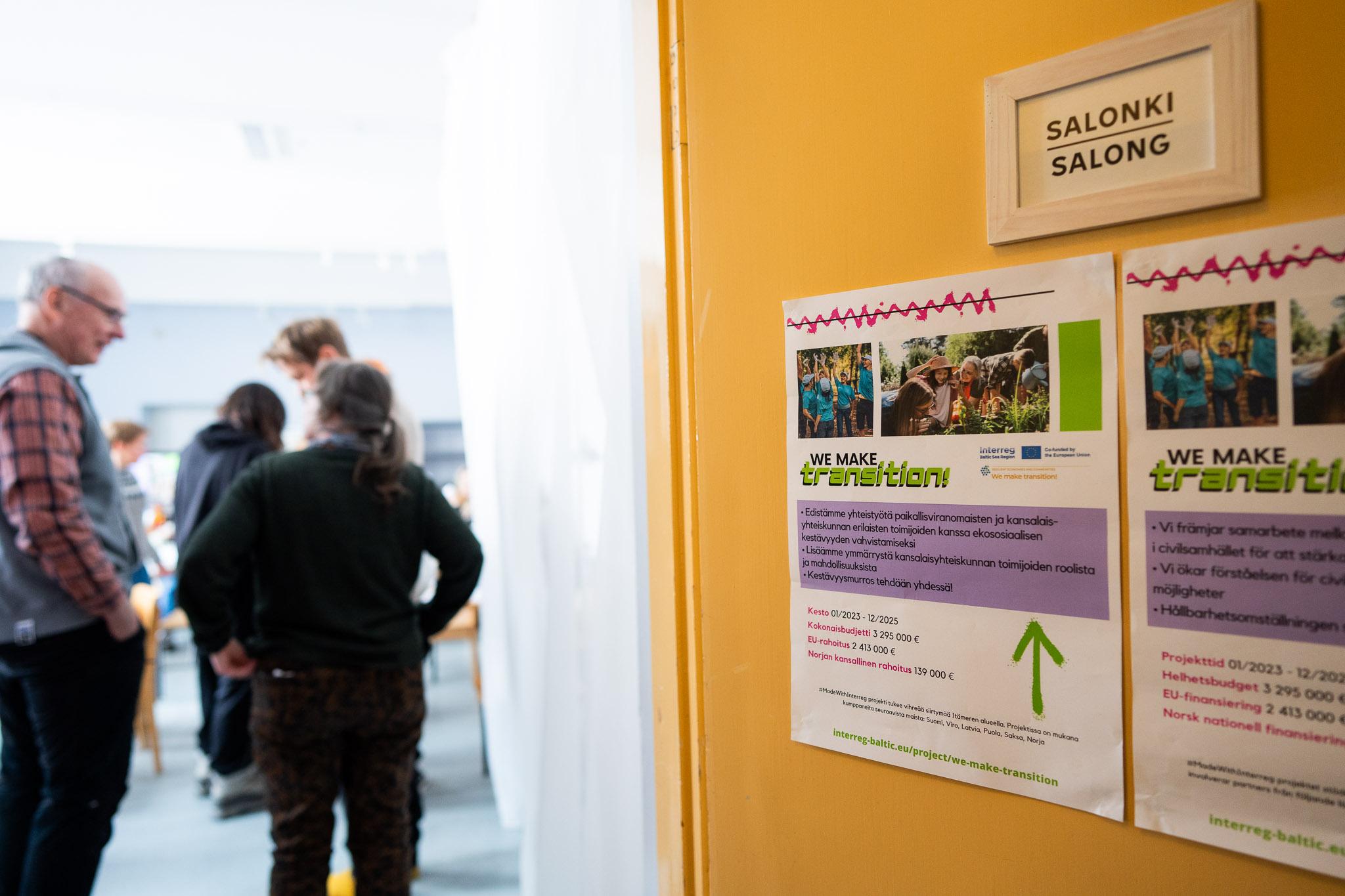 Ovi, jossa on kaksi värikästä julistetta. Ovesta aukeavassa huoneessa ihmisiä juttelemassa, tausta sumea.